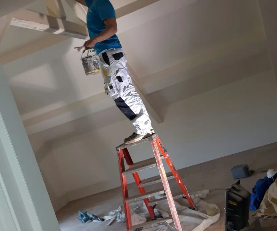 Schilder op trapladder lakt balken en plafond in een woning