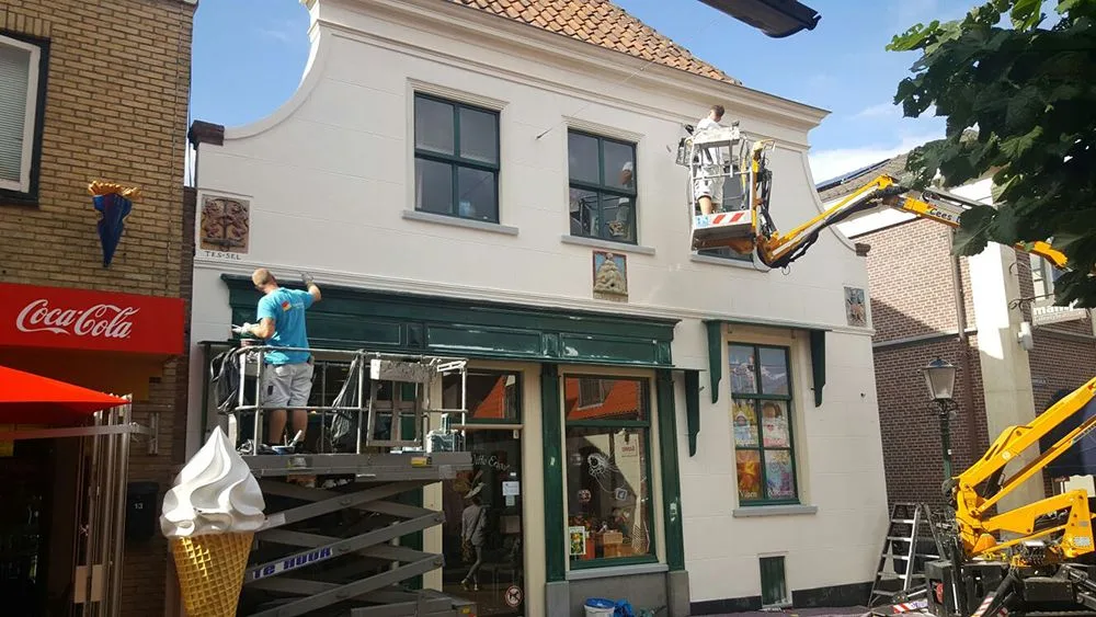 Schilders werken met een hoogwerker aan een wit winkelpand in Den Burg
