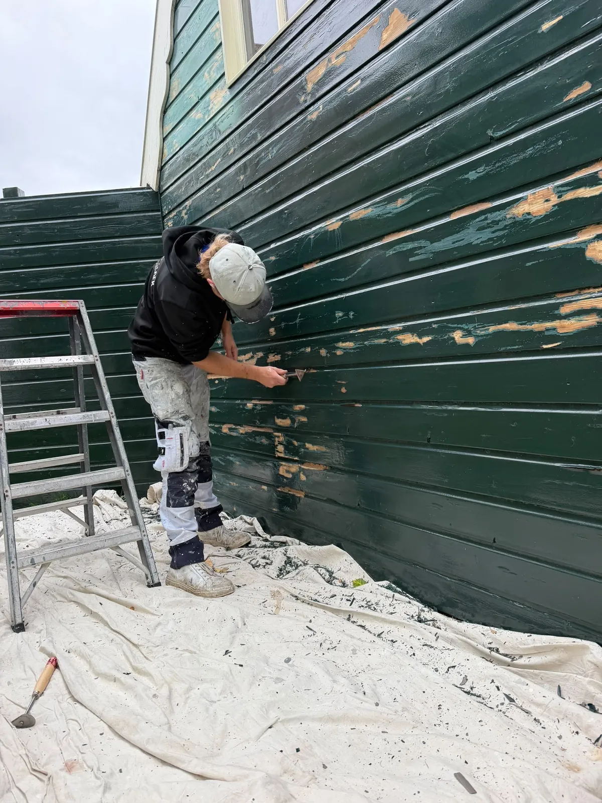 Schilder schuurt de oude verf van een donkergroene houten gevel