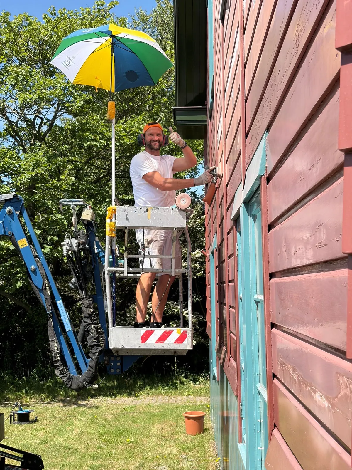 Schilder in een hoogwerker onder een parasol naast een rode houten gevel