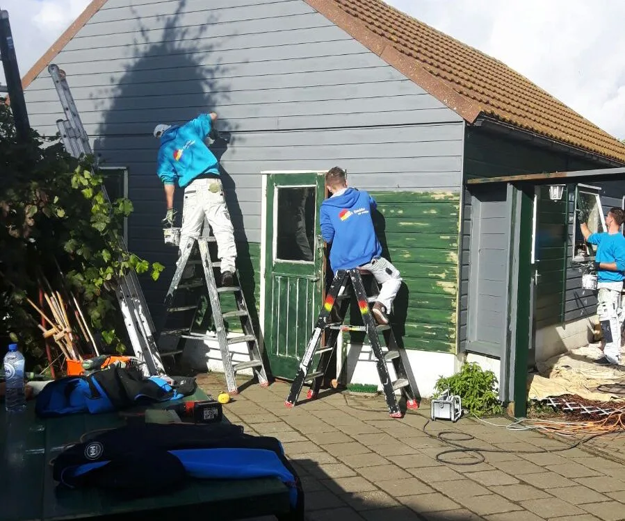 Drie schilders op ladders schilderen een houten schuur groen